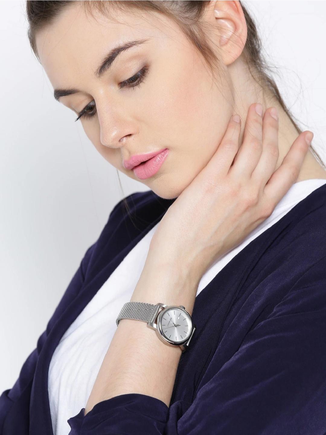 A woman wearing a silver mesh-strand analog watch with a silver dial and a dark navy velvet cardigan over a white top, posing with her hand on her neck against a light gray background.