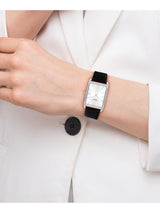 A woman wearing a minimalist rectangular analog watch with a silver-toned case, white dial, and black strap, shown against a white blazer.
