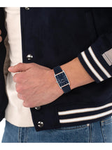 A close-up of a person wearing a rectangular analog watch with a dark navy blue strap and matching dial, styled with a navy and white varsity jacket and blue jeans.