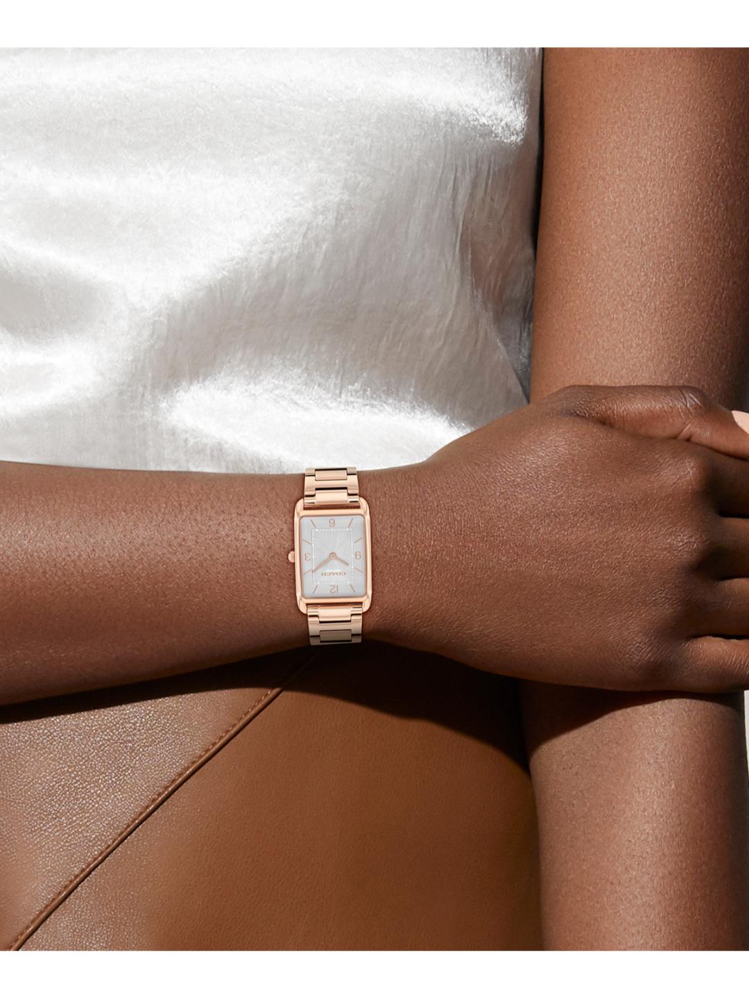 A close-up of a woman's wrist wearing a rectangular rose gold watch with a white dial, resting against a white satin fabric and brown leather background.