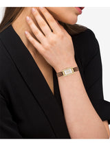 A close-up of a woman wearing a slim rectangular gold-tone watch with a textured brown strap, styled with a black blazer against a light gray background.