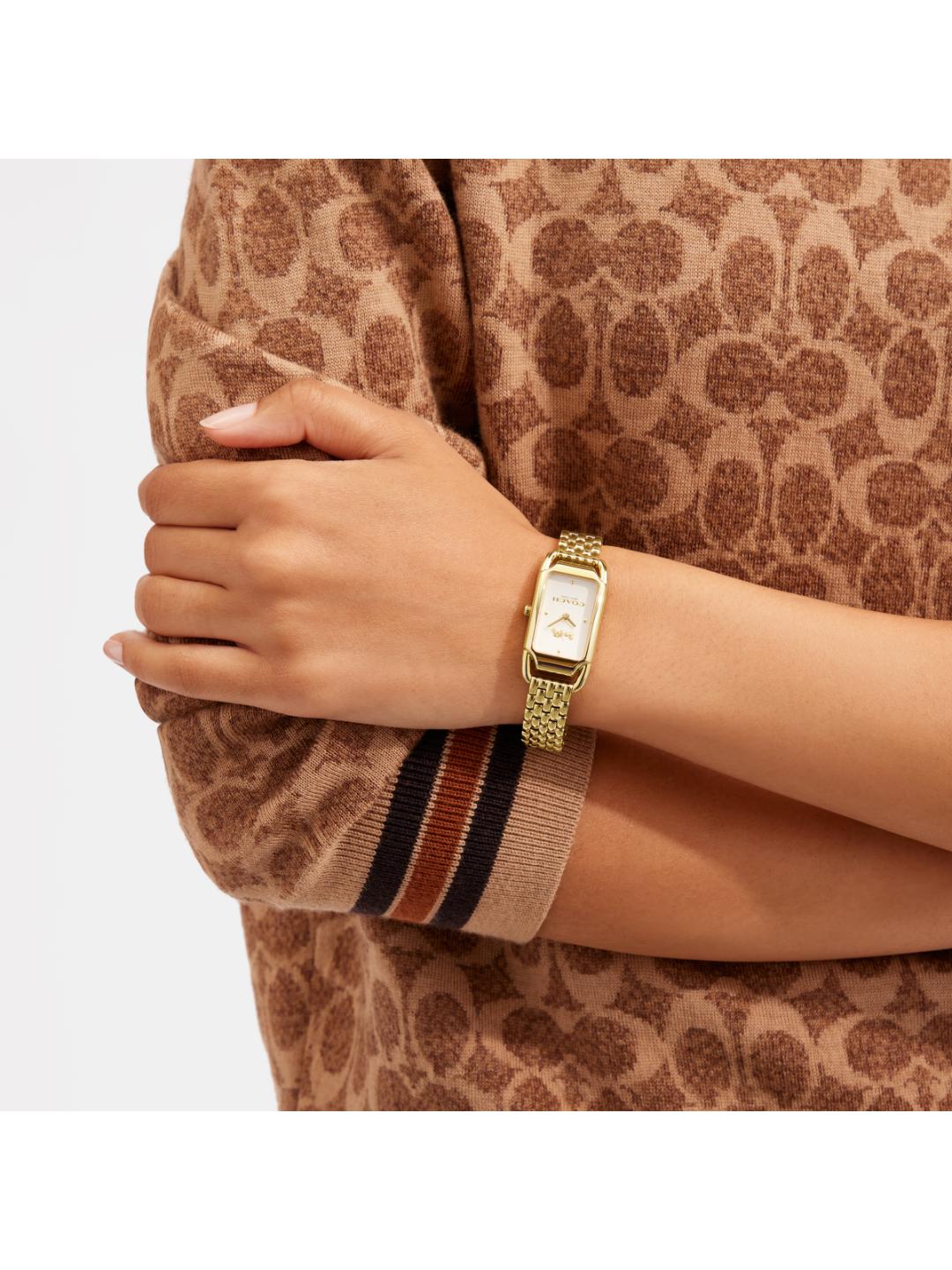 A close-up of a woman's arm wearing a gold-tone rectangular watch with a mesh band, styled with a brown and beige patterned knit sweater.