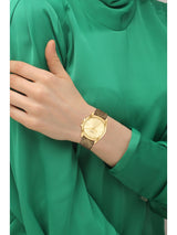 A gold-tone analog watch with a textured mesh strap worn on a person's wrist, paired with a shiny green satin turtleneck top.