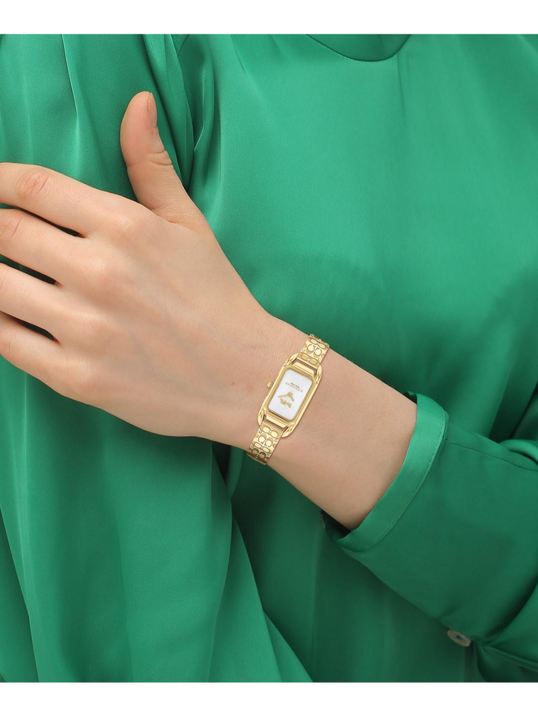 A gold-tone rectangular watch with a white dial and textured link bracelet, shown worn on a person's wrist against a green satin blouse.