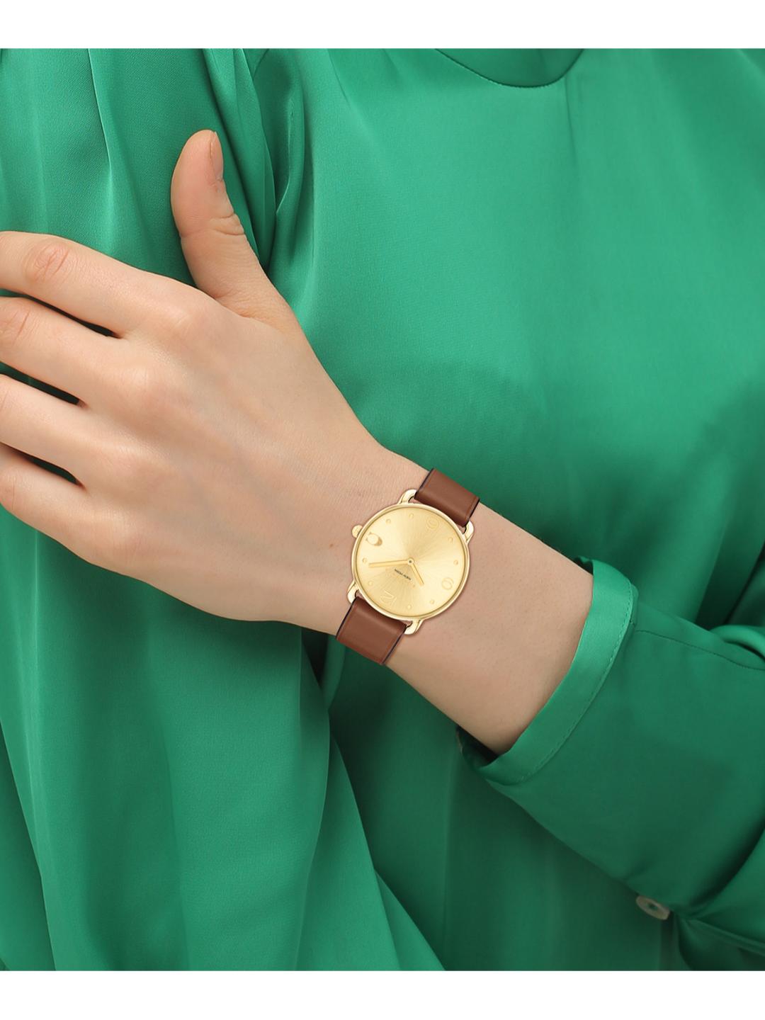 A close-up of a person's hand and wrist wearing a slim analog watch with a round gold-tone dial and brown leather strap, set against a bright green satin blouse.