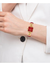 A close-up of a woman's wrist wearing a gold-tone watch with a red dial and red strap, styled with a white blazer.
