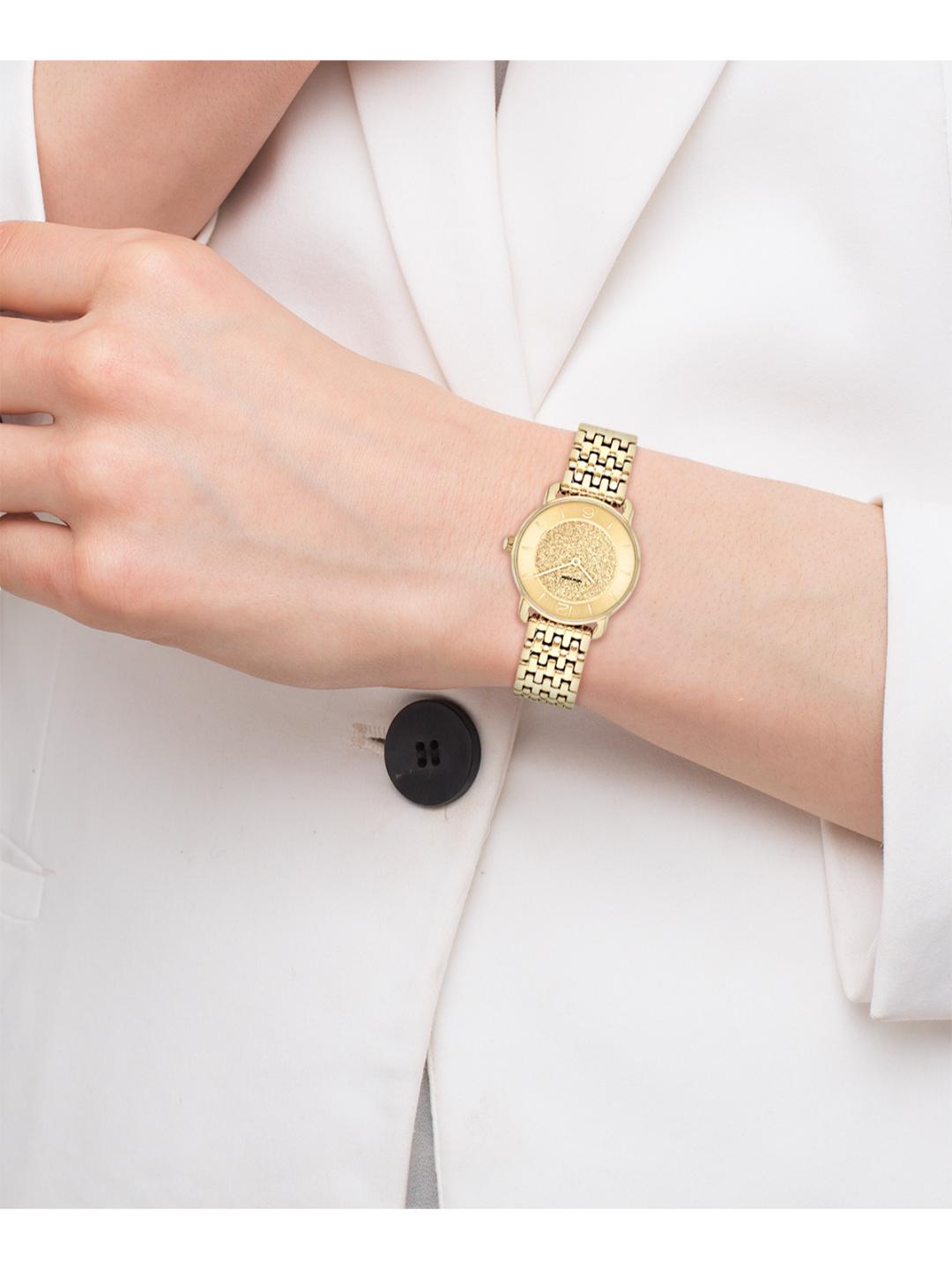 A gold-tone women's watch with a textured dial and matching link bracelet, shown worn on a person's wrist over a white blazer.