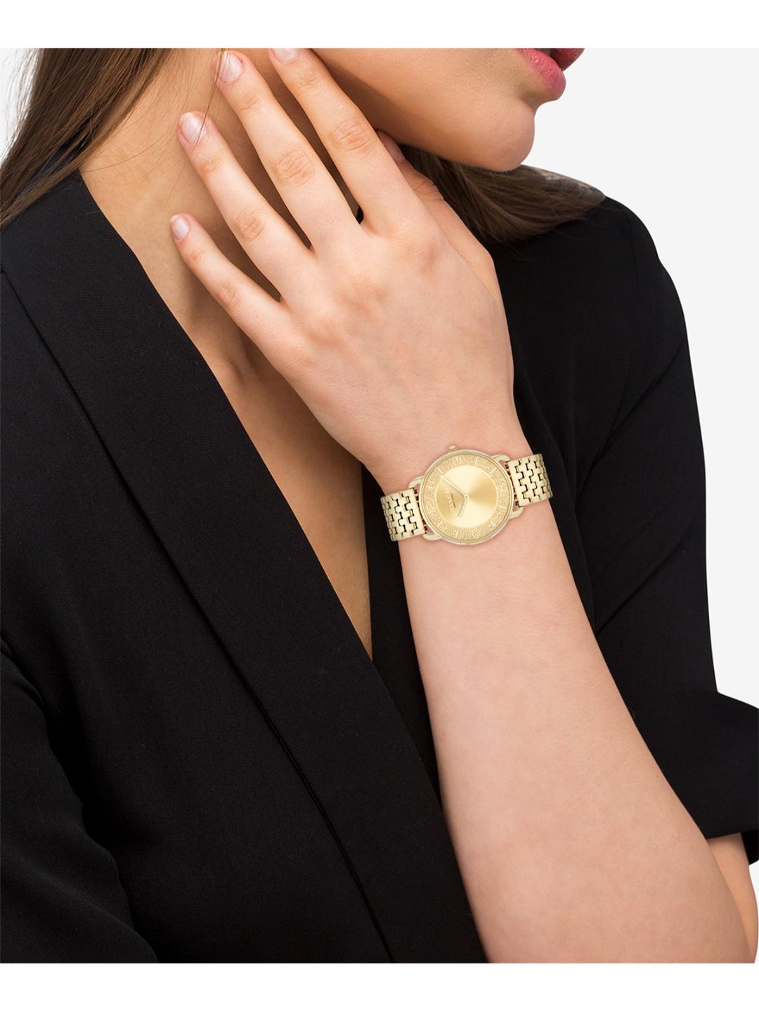 A close-up of a woman wearing a gold-tone watch with a textured metal link band and a round gold dial, paired with a black blazer.