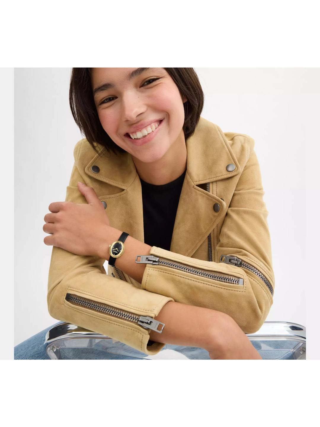 A smiling woman wearing a tan suede moto jacket with silver zipper details, a black top, and a watch, leaning on a clear plastic stool against a light gray background.
