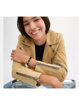 A smiling woman wearing a tan suede moto jacket with silver zipper details, a black top, and a watch, leaning on a clear plastic stool against a light gray background.