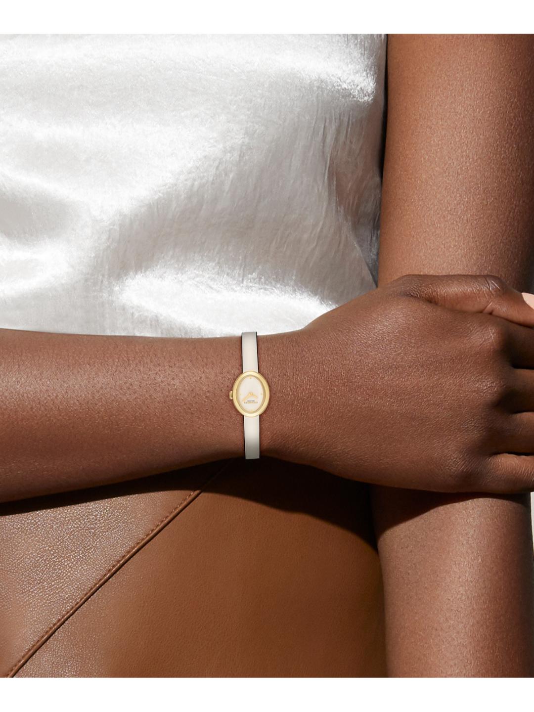 A close-up of a person's wrist wearing a delicate white leather bangle bracelet with a small oval gold-toned medallion.