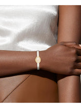 A close-up of a person's wrist wearing a delicate white leather bangle bracelet with a small oval gold-toned medallion.