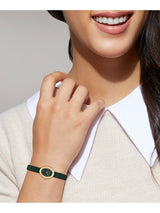 A close-up of a woman wearing a slim dark green leather wristband with a gold-tone oval bezel and a matching green dial.