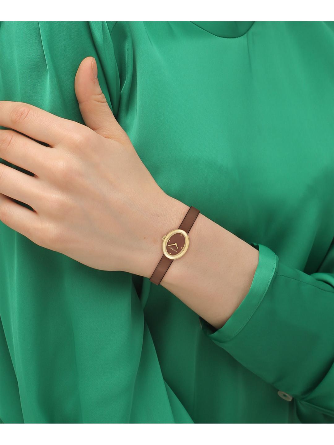 A close-up of a person's hand and wrist wearing a slim brown leather bracelet watch with a gold-tone round case and minimalist dial, set against a vibrant green satin blouse.