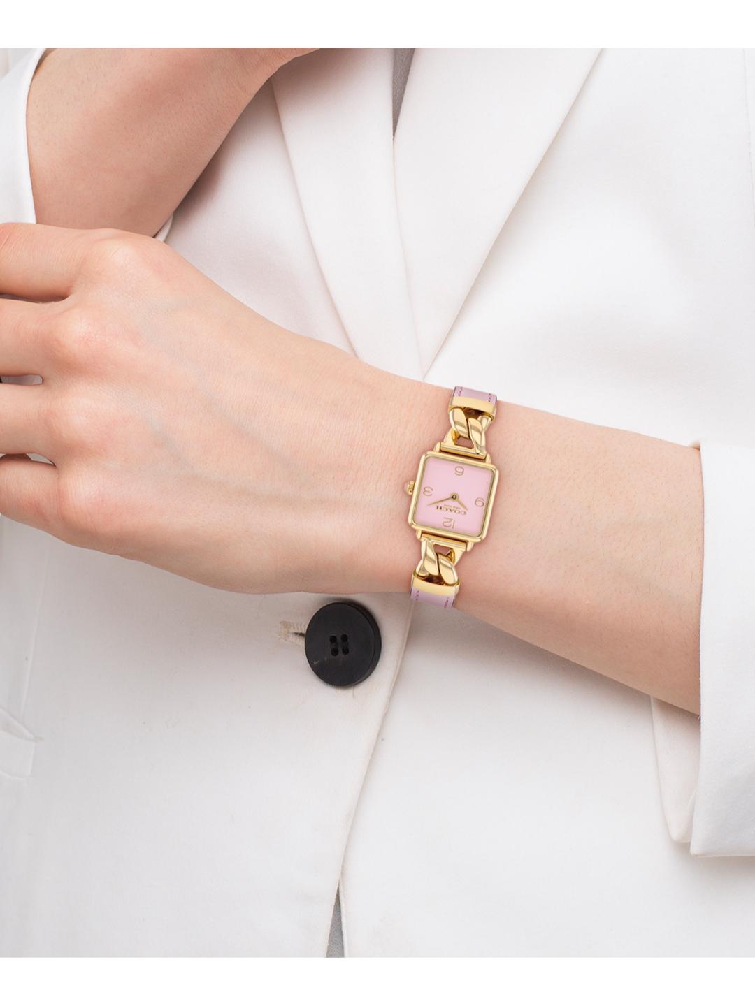 A gold-tone watch with a pink dial and matching pink strap, worn on a model's wrist over a white blazer.