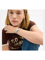 A woman wearing a silver-toned analog watch with a light blue mesh strap and a white dial, styled with a brown ringer t-shirt and blue jeans against a plain white background.