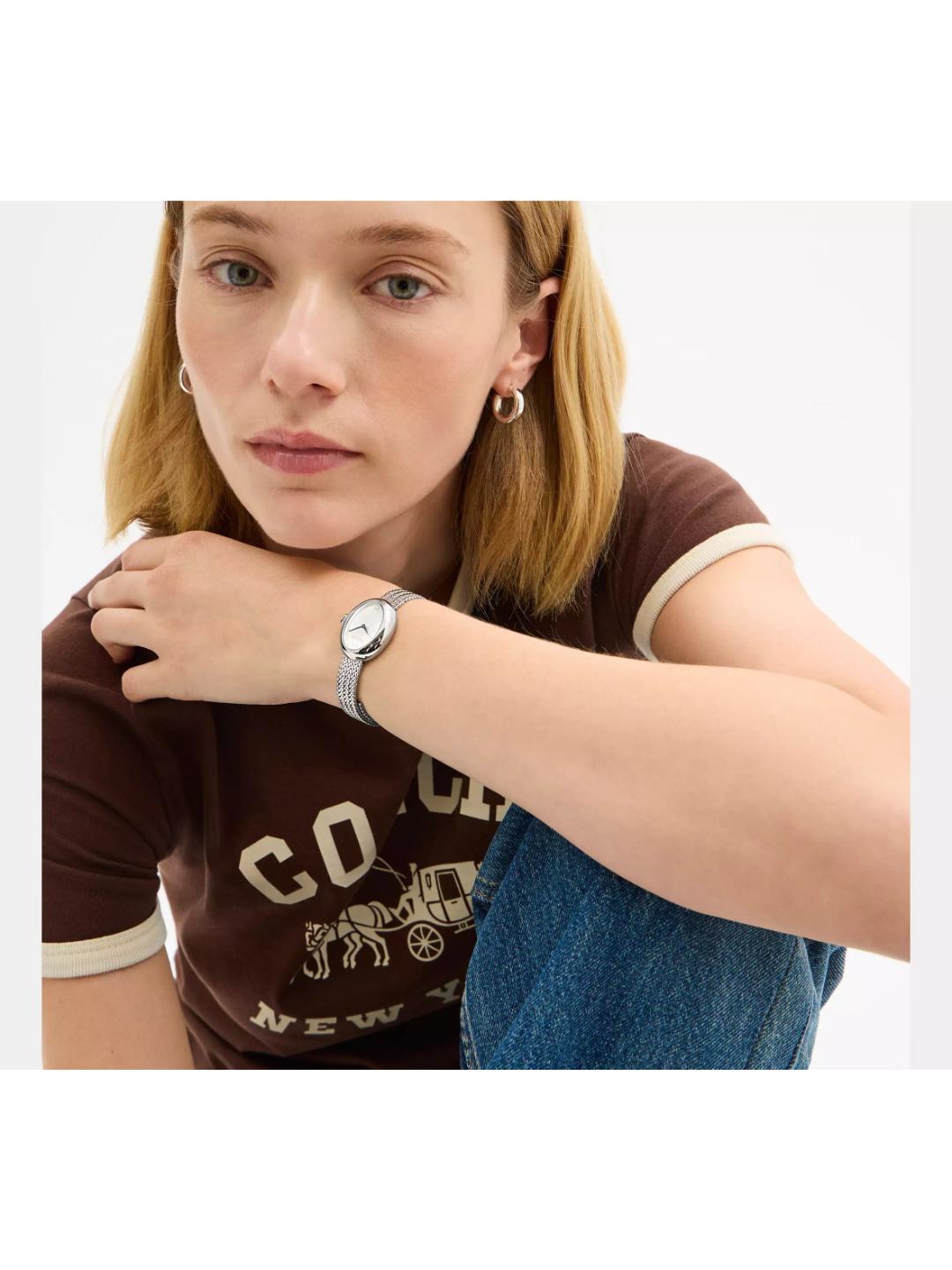 A woman wearing a silver-toned analog watch with a light blue mesh strap and a white dial, styled with a brown ringer t-shirt and blue jeans against a plain white background.