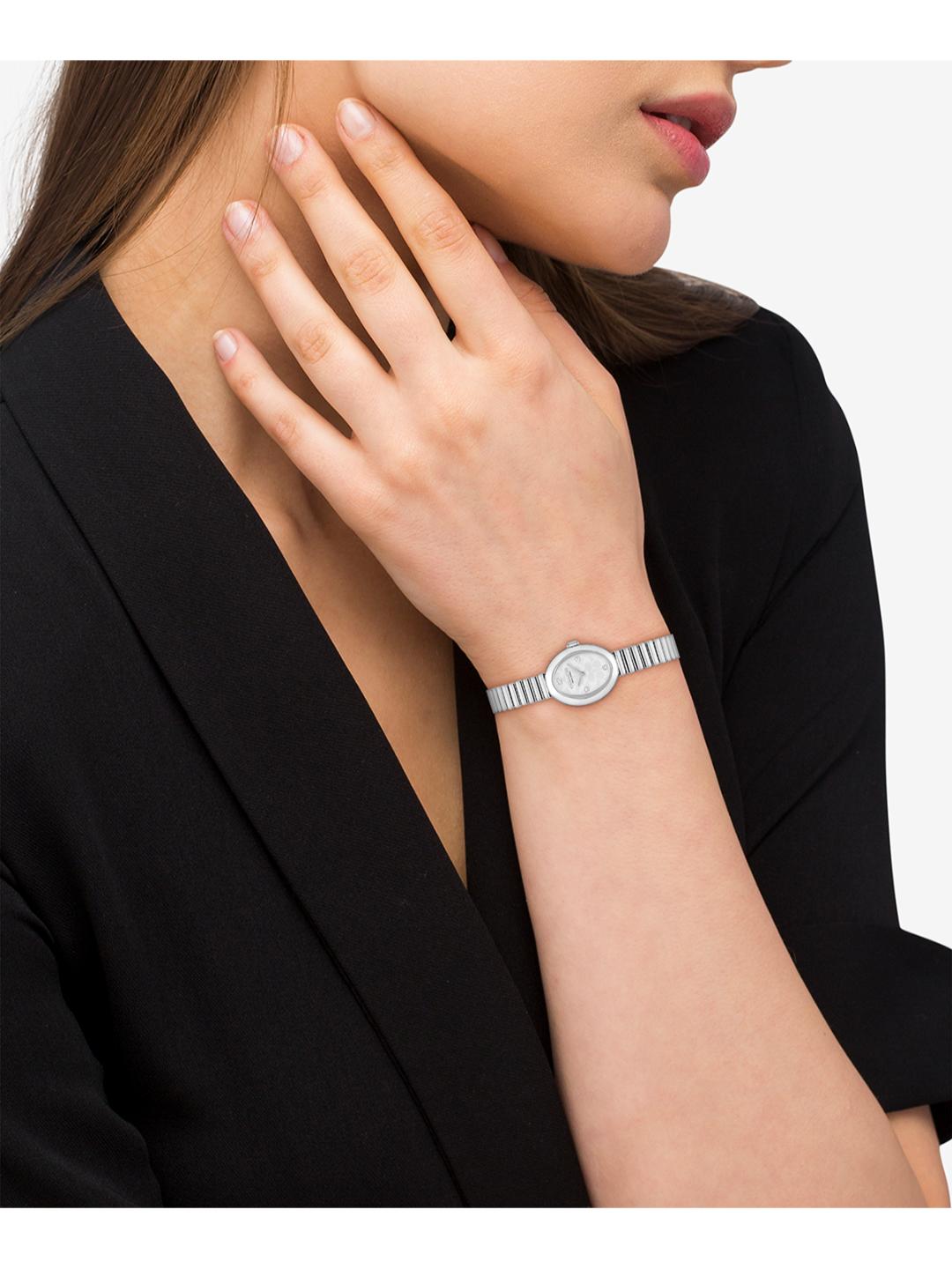 A close-up of a woman wearing a slim silver-tone watch with a matching metal link bracelet, paired with a black blazer against a light gray background.