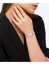 A close-up of a woman wearing a slim silver-tone watch with a matching metal link bracelet, paired with a black blazer against a light gray background.