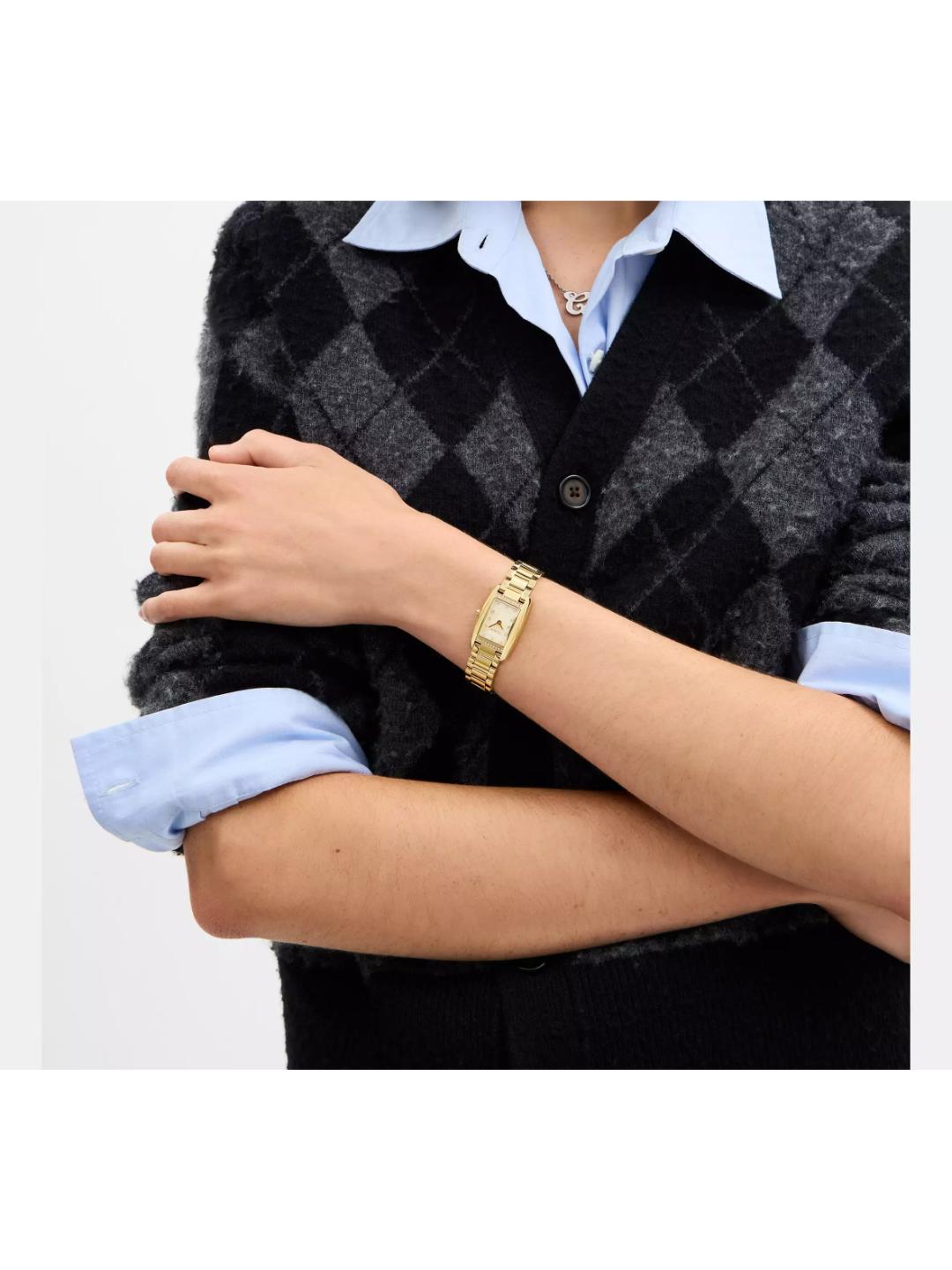 A close-up of a person wearing a gold-tone rectangular watch with a gold-tone link bracelet, styled with a black and gray argyle cardigan over a light blue collared shirt.