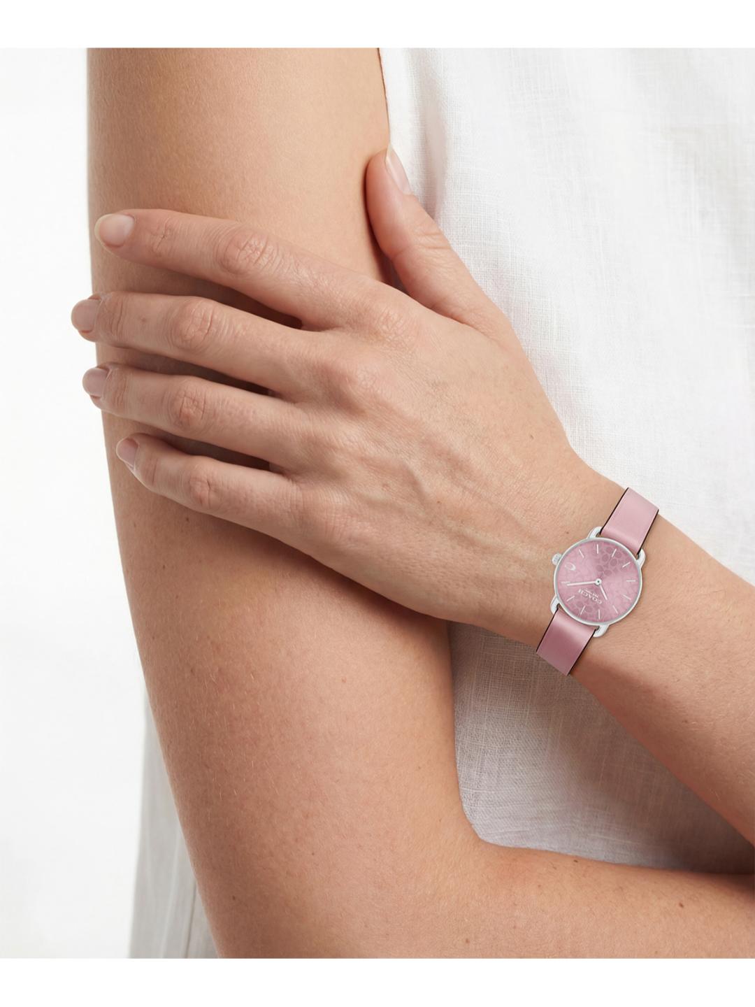 A close-up of a model's arm and hand wearing a slim pink watch with a silver-toned bezel and matching pink strap.