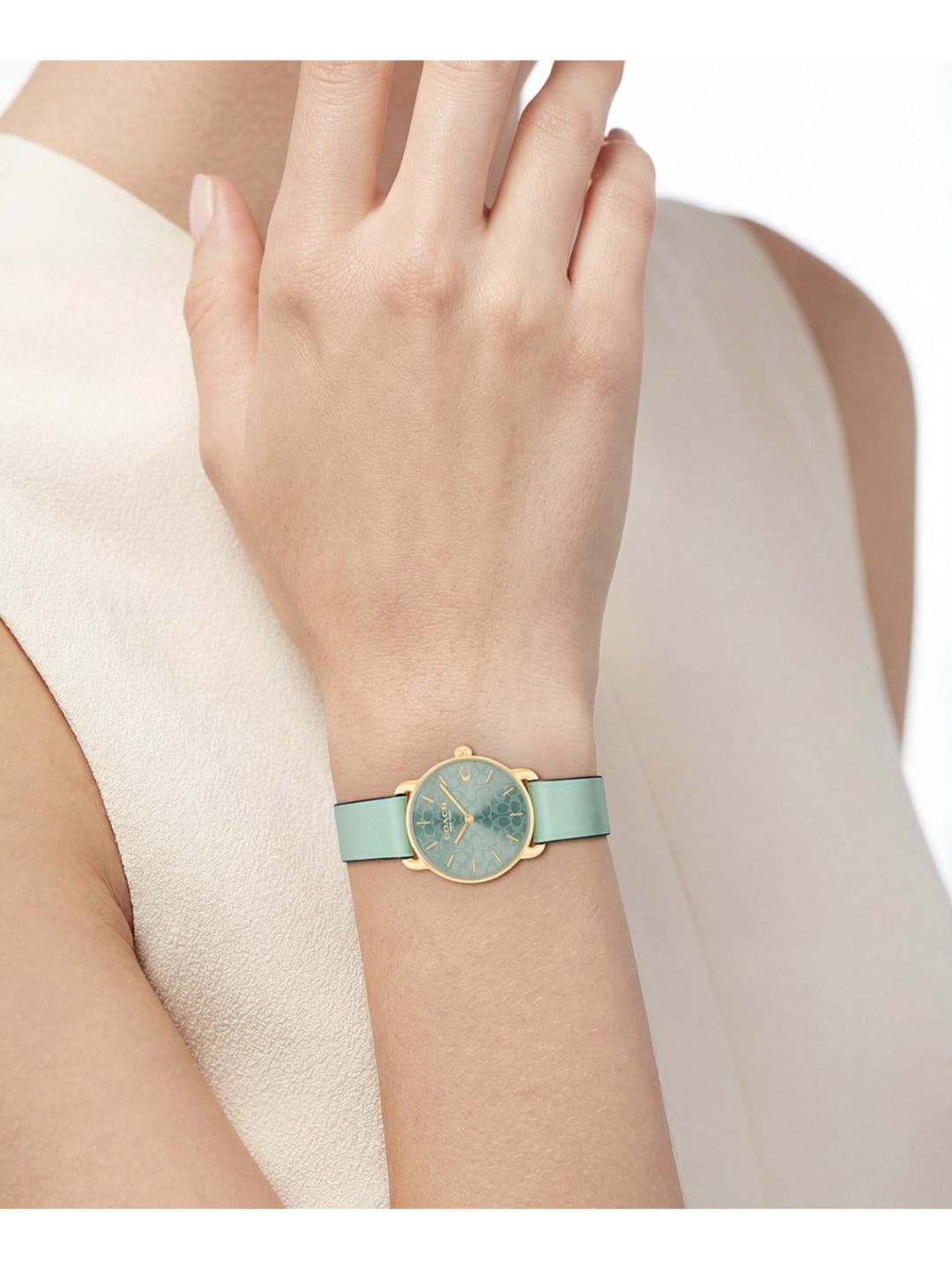 A close-up of a woman's wrist wearing a minimalist watch with a light mint green strap, a gold-tone case, and a textured dial.