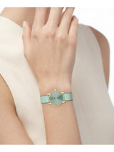A close-up of a woman's wrist wearing a minimalist watch with a light mint green strap, a gold-tone case, and a textured dial.