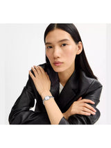 A woman with long black hair wearing a black leather jacket and a silver-toned watch with a white dial, posed against a plain white studio background.