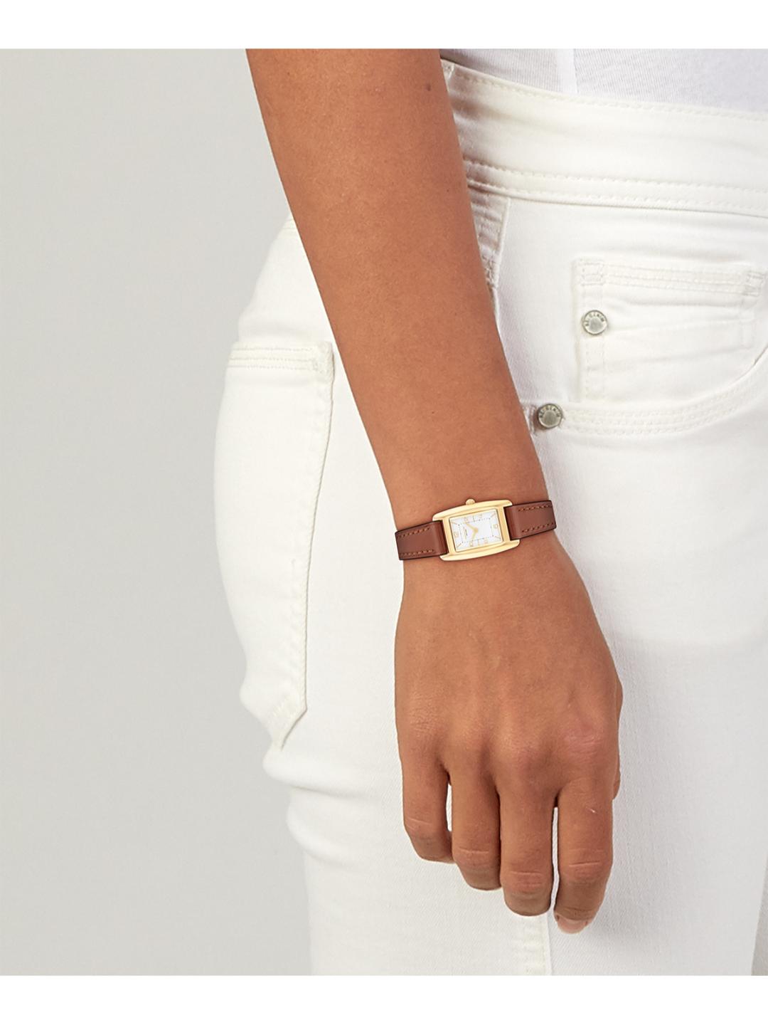 A close-up of a person's wrist wearing a small rectangular watch with a brown leather strap and a gold-tone case, paired with white jeans against a light gray background.