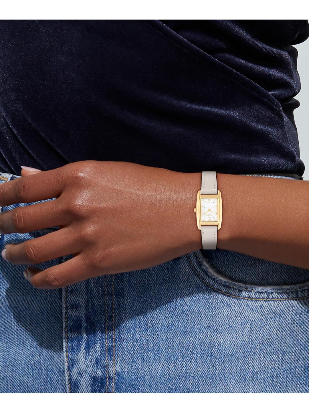 A close-up of a woman's wrist wearing a slim rectangular watch with a light beige strap and gold-tone case, paired with a dark navy velvet top and blue jeans.