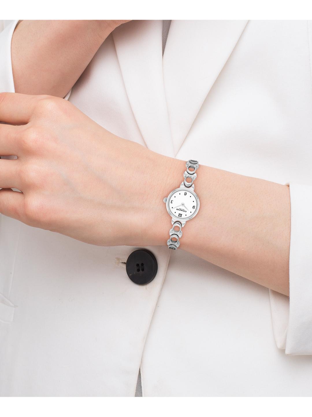 A silver-tone women's watch with a white dial and a distinctive segmented link bracelet, shown worn on a person's wrist. The person is wearing a white blazer, and the watch is positioned over the white button of the blazer.