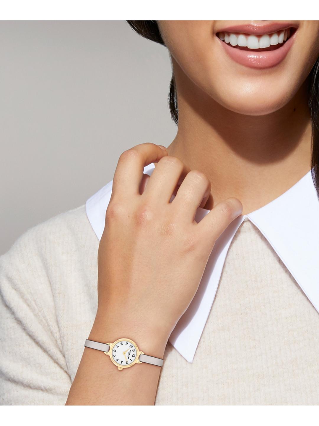 A close-up of a woman wearing a slim gold-tone watch with a white strap and white dial, paired with a white collared shirt and beige knit top.