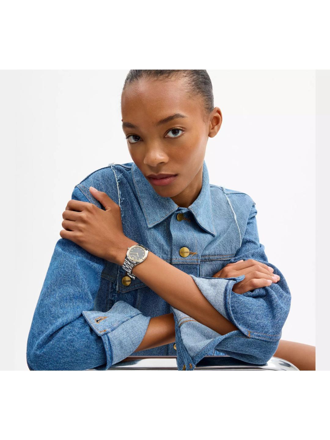 A model wearing a blue denim shirt and a silver-toned watch with a round bezel and matching metal bracelet, posed against a clean white background.