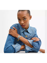 A model wearing a blue denim shirt and a silver-toned watch with a round bezel and matching metal bracelet, posed against a clean white background.