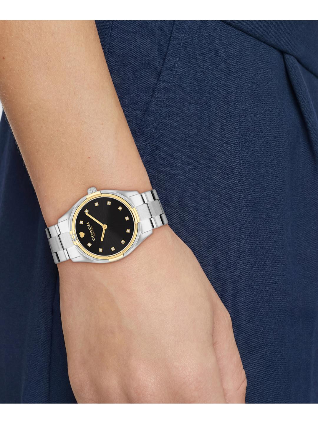 A close-up of a woman's wrist wearing a Movado watch with a black dial, gold-tone details, and a silver and gold two-tone metal bracelet, set against a navy blue dress.