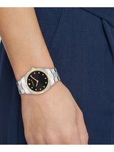 A close-up of a woman's wrist wearing a Movado watch with a black dial, gold-tone details, and a silver and gold two-tone metal bracelet, set against a navy blue dress.