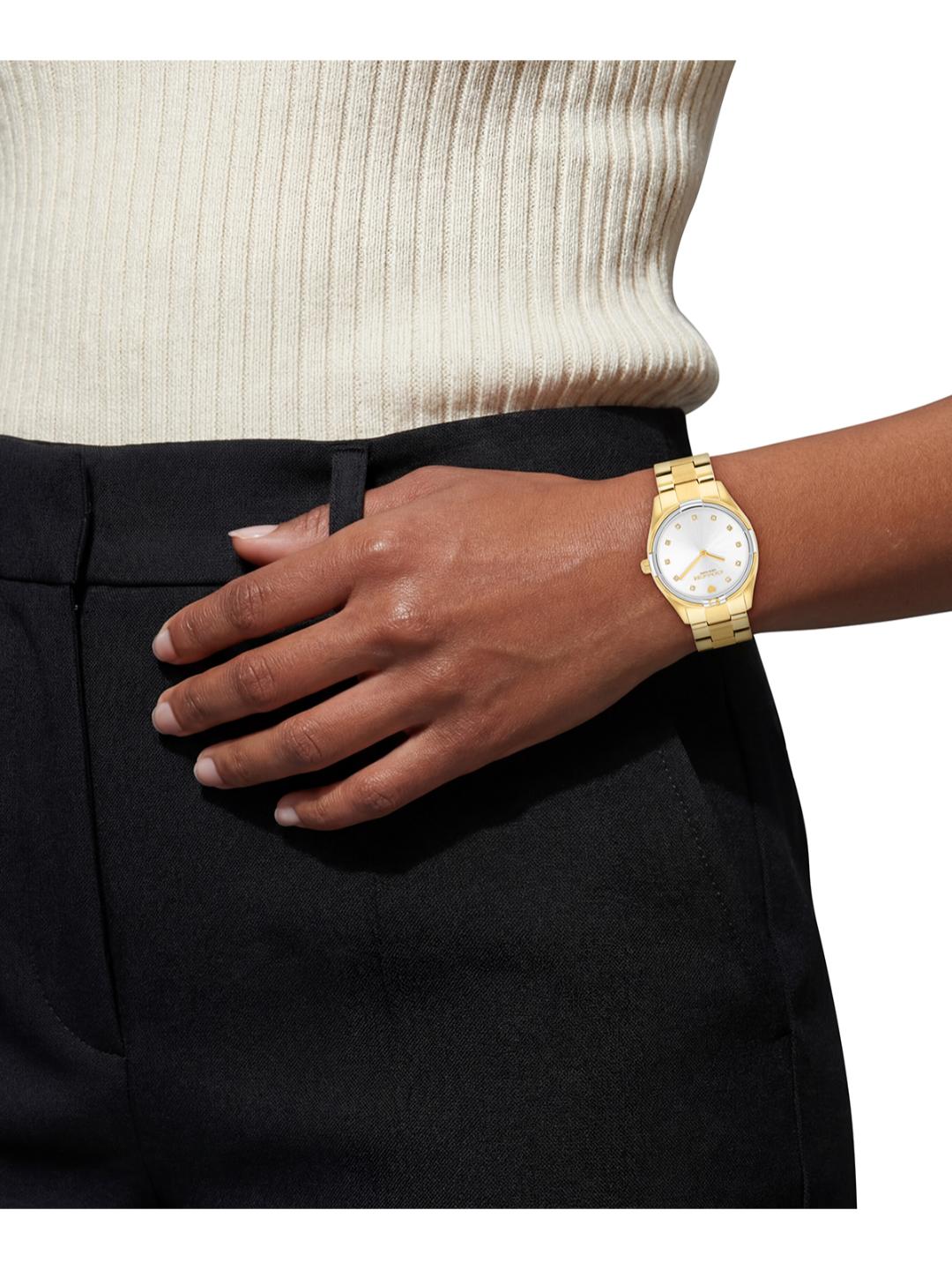 A gold-tone metal watch with a white dial and crystal case, worn on a person's wrist. The person is wearing a cream ribbed knit top and black trousers, with their hand resting on their thigh against a plain white background.