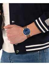 A wristwatch with a blue dial and gold-tone case worn on a person's wrist, shown under a navy varsity jacket with white stripes and blue jeans.