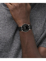 A close-up of a man's wrist wearing a Calvin Klein analog watch with a black dial, silver-tone case, and black strap, set against a gray heathered t-shirt.