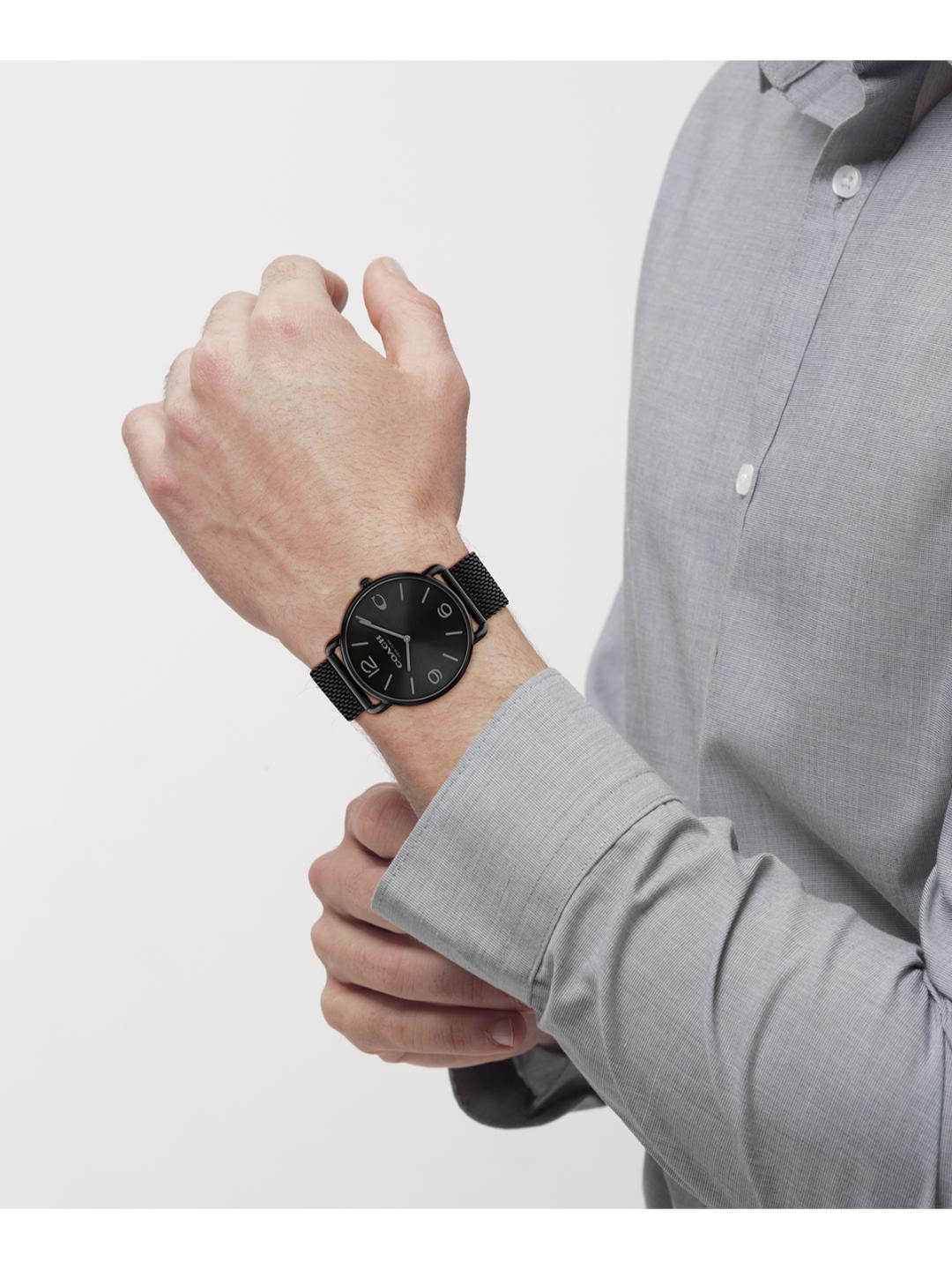 A man wearing a black dial watch with a black strap, shown from the chest down against a light gray background.