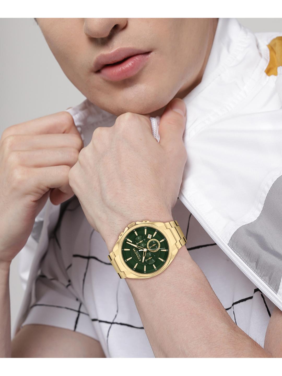 A gold-tone men's watch with a dark green dial and matching green bezel, worn on a model's wrist. The model is wearing a white and gray patterned shirt.
