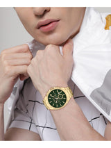 A gold-tone men's watch with a dark green dial and matching green bezel, worn on a model's wrist. The model is wearing a white and gray patterned shirt.