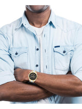 A man wearing a light blue denim shirt and white t-shirt, with his arms crossed, showcasing a gold-tone wristwatch with a black dial.