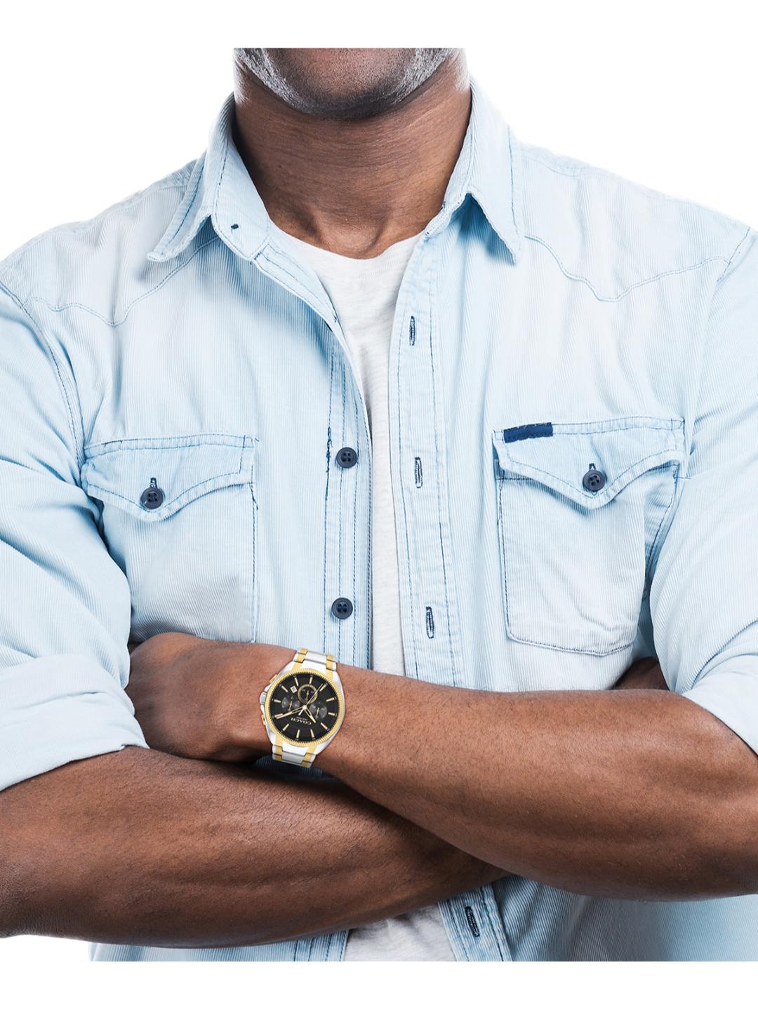 A man wearing a light blue denim shirt and white t-shirt, with his arms crossed, showcasing a gold-tone wristwatch with a black dial.