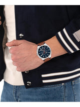 A men's wristwatch with a dark blue dial and silver-tone case, worn on a person's wrist. The person is wearing a navy blue varsity jacket with white stripes, a cream knit shirt, and blue jeans.