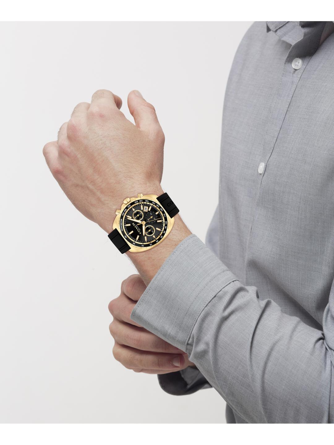 A man wearing a gold-tone chronograph watch with a black dial and black strap, shown from the chest down against a light gray studio background.