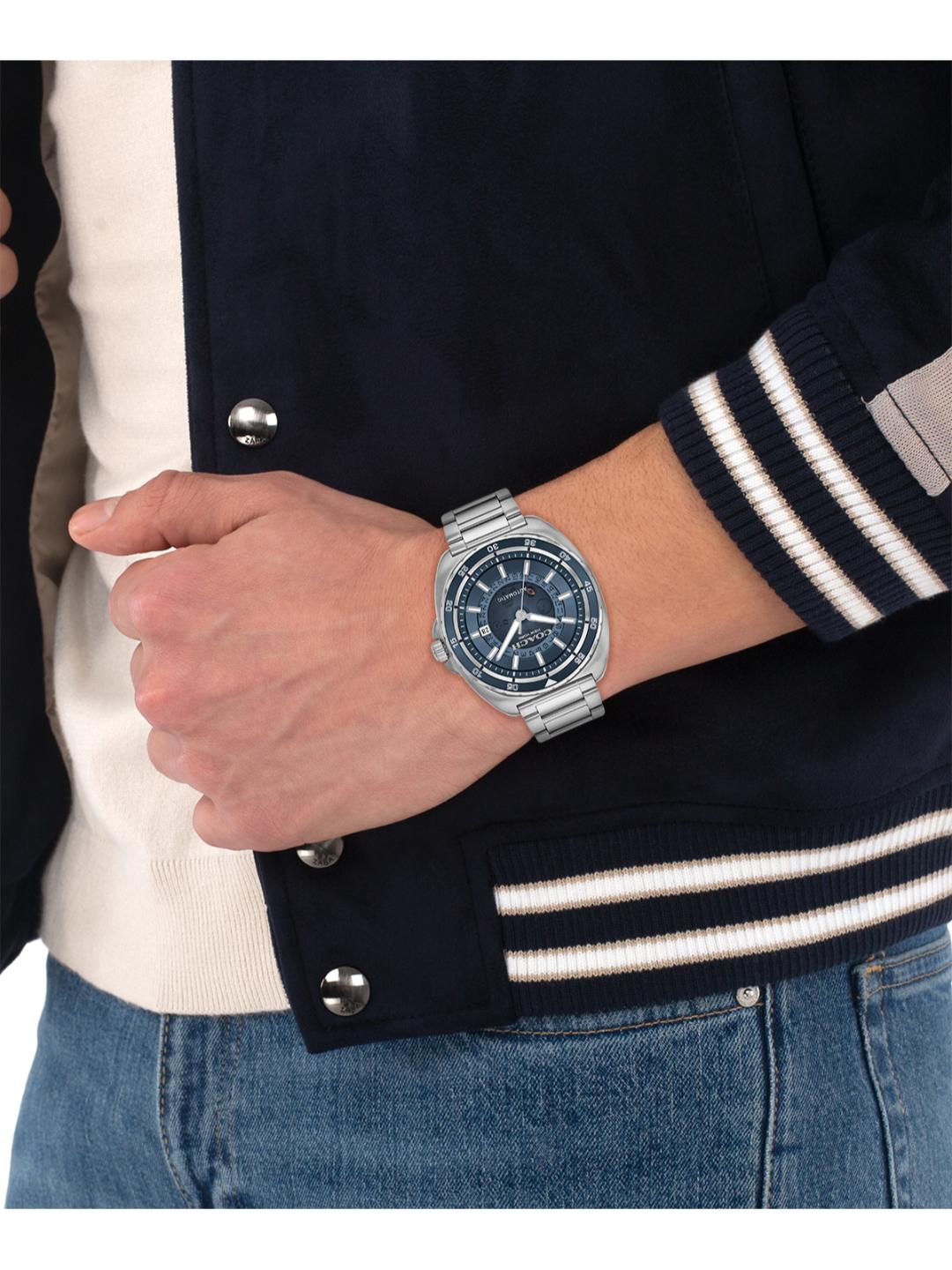 A silver stainless steel wristwatch with a blue dial and bezel, worn on a person's wrist. The person is wearing a navy blue varsity jacket with white stripes on the sleeves, a cream ribbed shirt, and blue jeans.