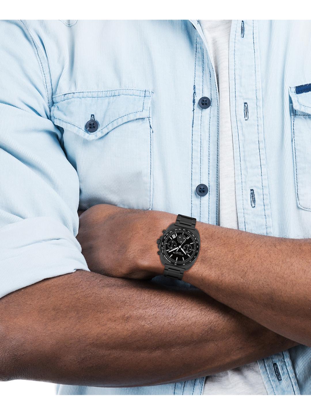A close-up of a man's crossed arms wearing a black multi-function wristwatch with a black strap, styled with a light blue denim shirt.