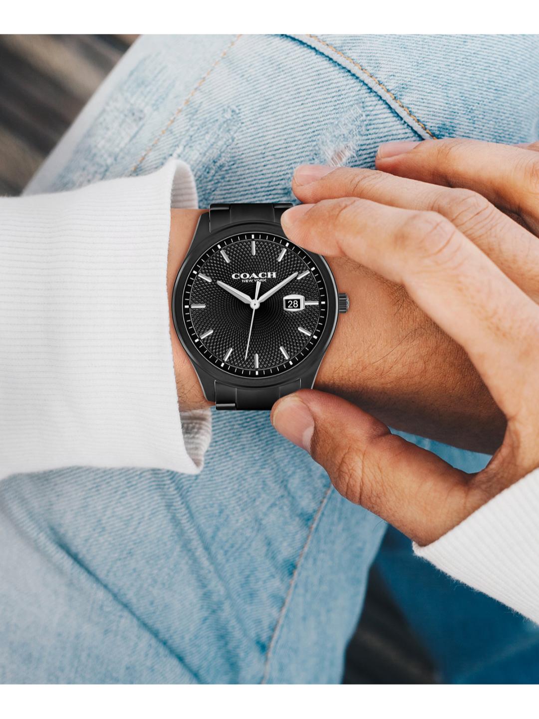 A close-up lifestyle shot of a person wearing a Coach black analog watch with a date window, styled with light blue jeans and a white ribbed knit sweater.