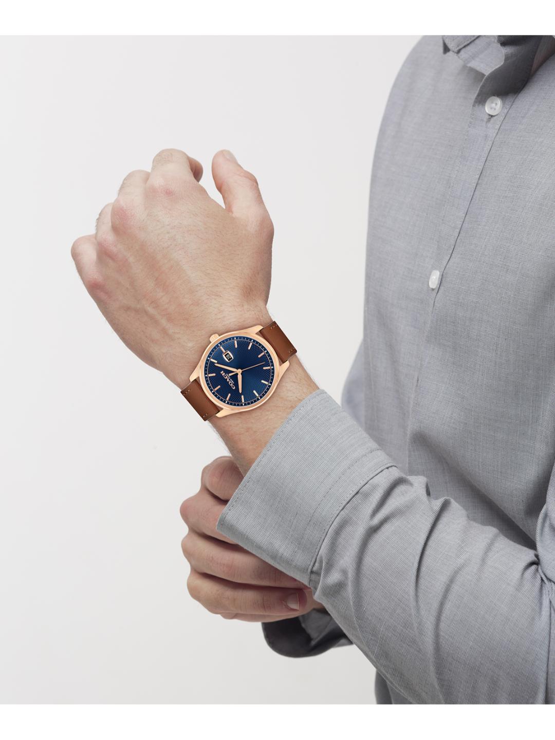 A man wearing a rose gold watch with a blue dial and brown leather strap, shown in a close-up lifestyle shot against a light gray background.